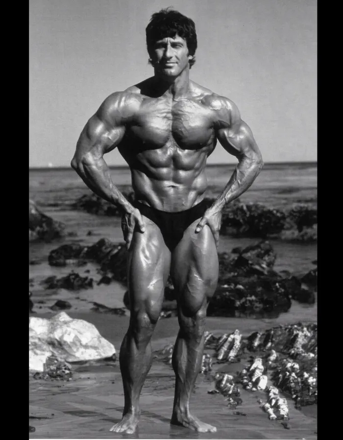 A magazine-quality most-muscular pose on the beach, rendered in stark black-and-white contrast with a direct, unflinching gaze.