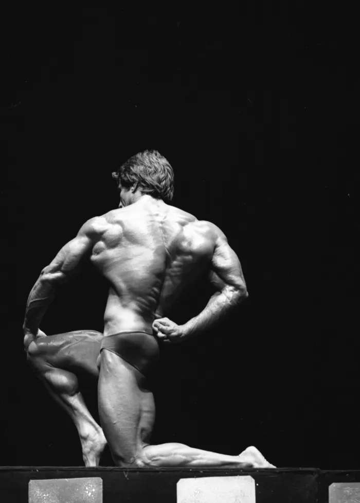 A kneeling back double-biceps pose shot from a striking low angle reveals full back detail.