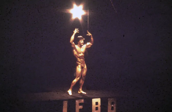 A triumphant victory moment atop the IFBB podium, arms raised in a starburst spotlight.