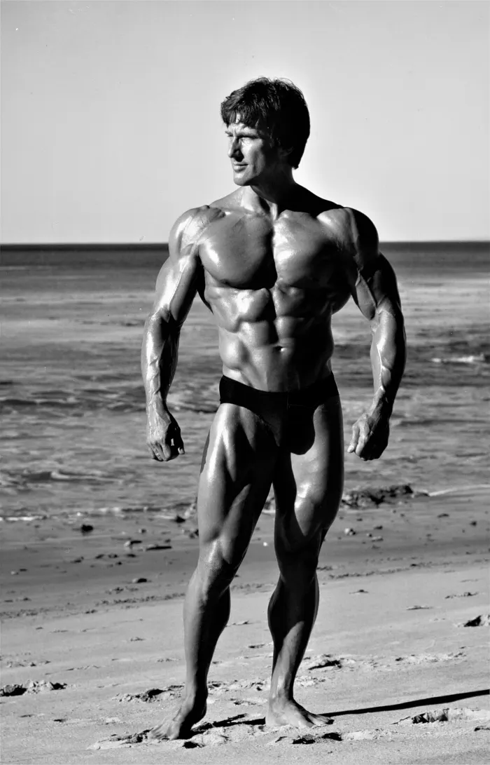 A medium-format full-body beach pose with dramatic backlighting and ocean backdrop.