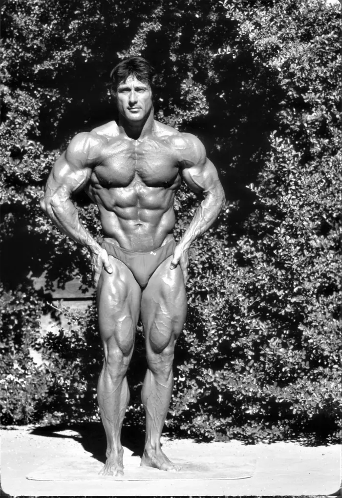 A relaxed full-front stance against foliage, exceptional muscle definition in the classic Zane physique showcase.