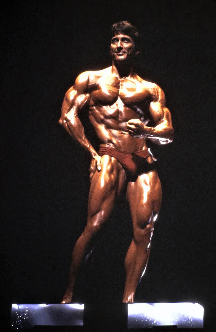 A dramatic spotlit stage pose in red posing trunks, genuine competition atmosphere.