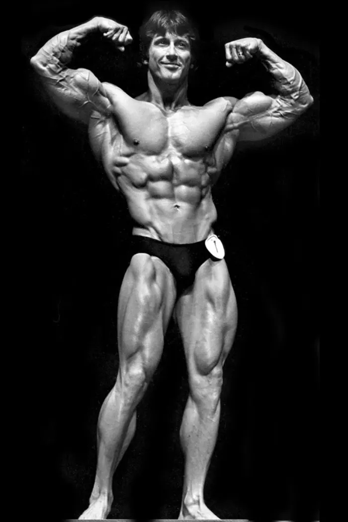 An iconic vintage black-and-white front double-biceps pose with contest number visible, peak 1977 Mr. Olympia-era drama.