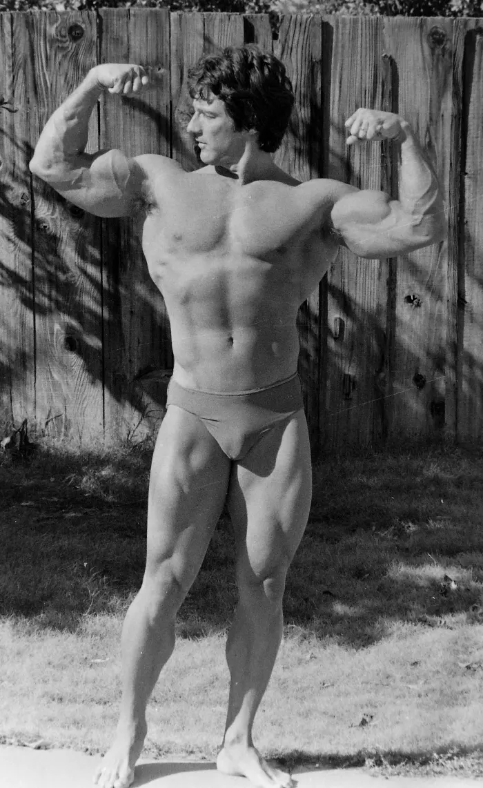 A dynamic rear double-biceps pose from a backyard photo shoot, well-lit black-and-white film showing clear back detail.