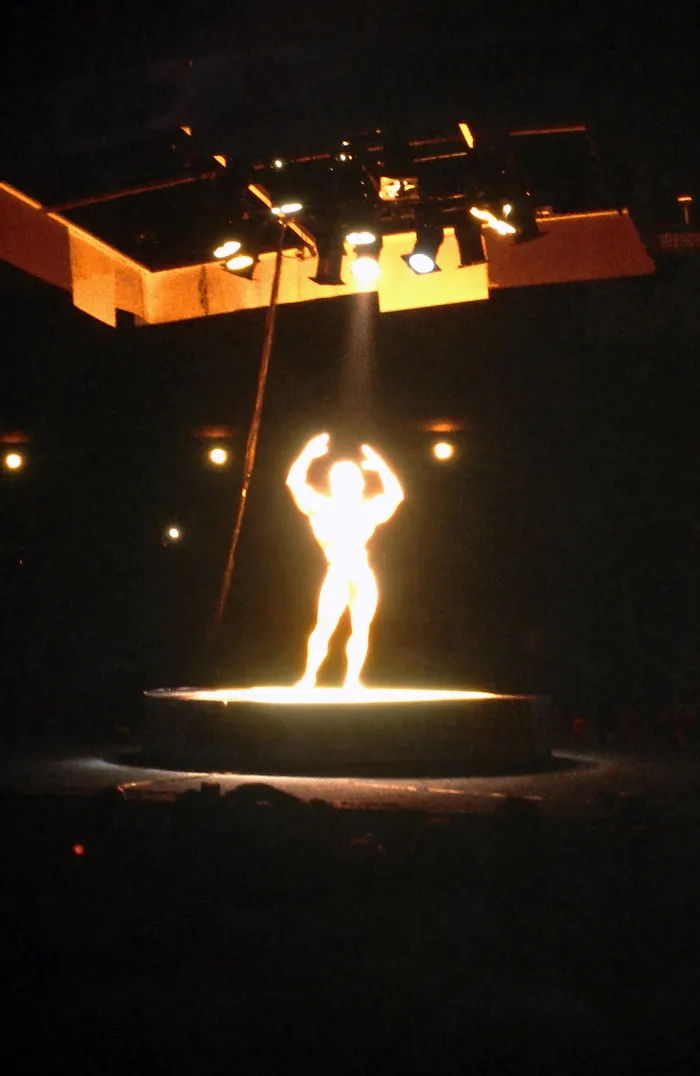 A backlit gold silhouette front double-biceps pose on an Olympia-style stage, magazine-cover drama.