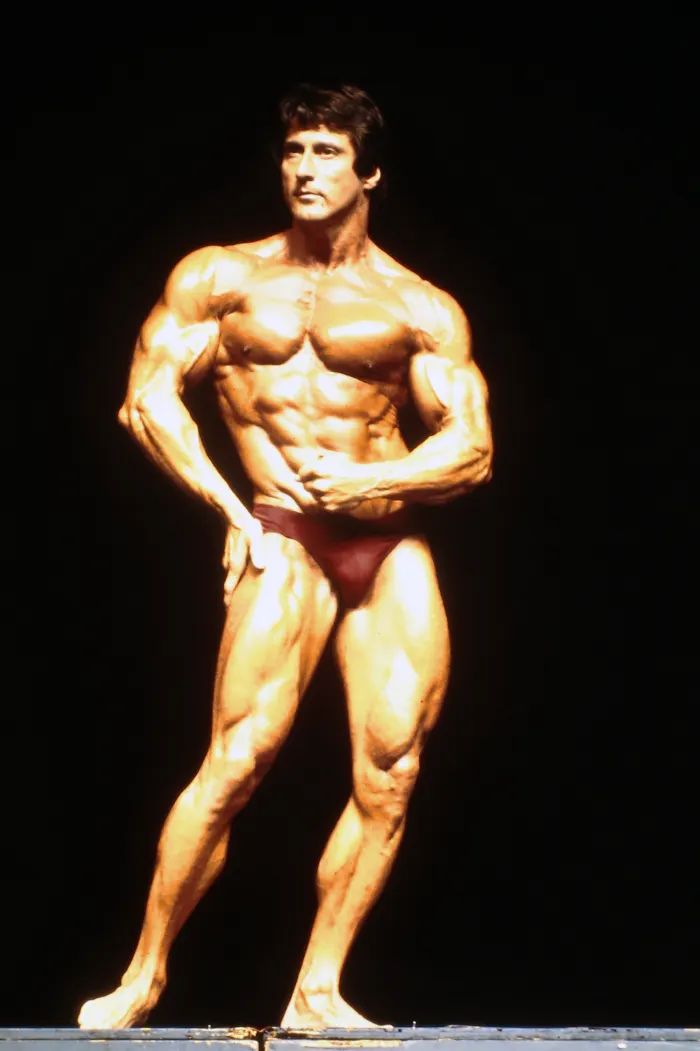 An oiled physique in a front lat-spread on ocean tide rocks at golden light, peak-condition abs and vascularity.