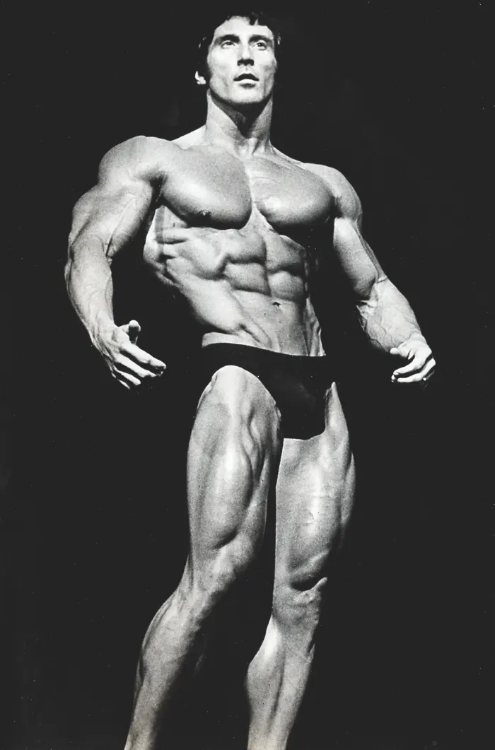 A dramatic studio-lit black-and-white lat-spread pose on pure black background, museum-quality contrast and intense gaze.
