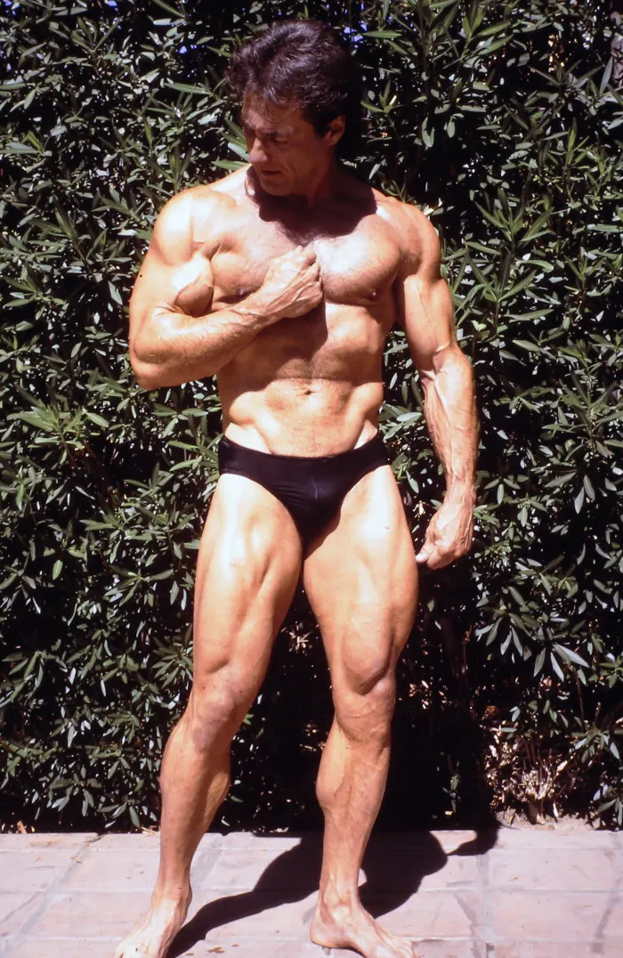 A genuine IFBB stage shot with banner and crowd visible, gold spotlighting, a powerful front double-biceps stare.