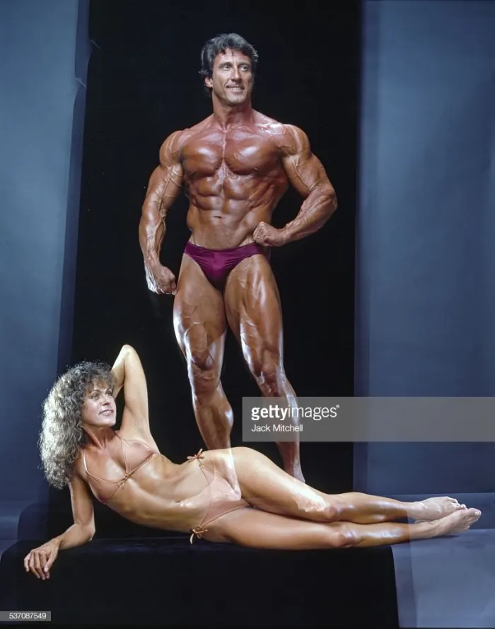 A professional studio portrait with dramatic blue backdrop and glamour lighting, a full-length double-biceps pose.
