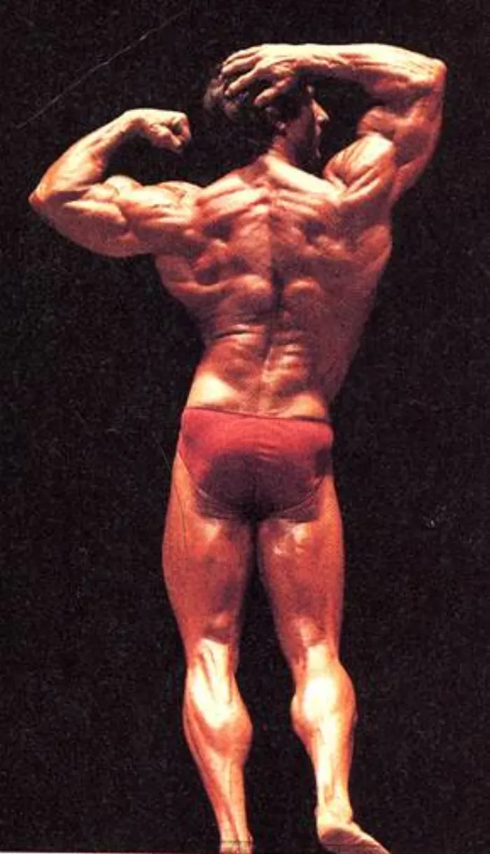 A historically significant Mr. Olympia-era color stage shot, red trunks against an outdoor desert backdrop.