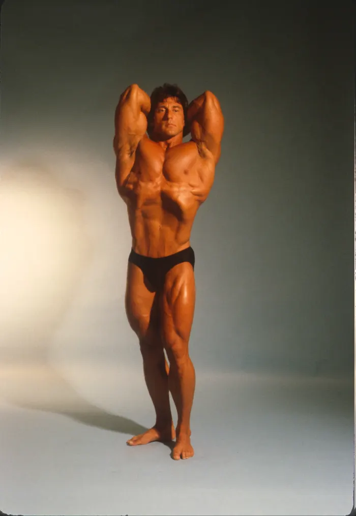 A polished full-color studio rendition of the signature vacuum double-biceps pose, professional lighting and tan.